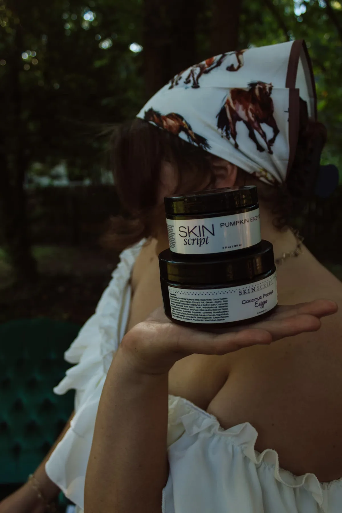 Two stacked amber skincare jars held out on an open palm, horse-print scarf in the background